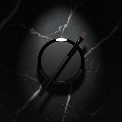 Overhead flat lay of closed matte black eyeshadow compact with black brush on dark marble
