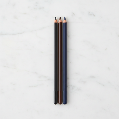 Three matte wooden eyeliner pencils in black, brown and navy standing side by side on white marble